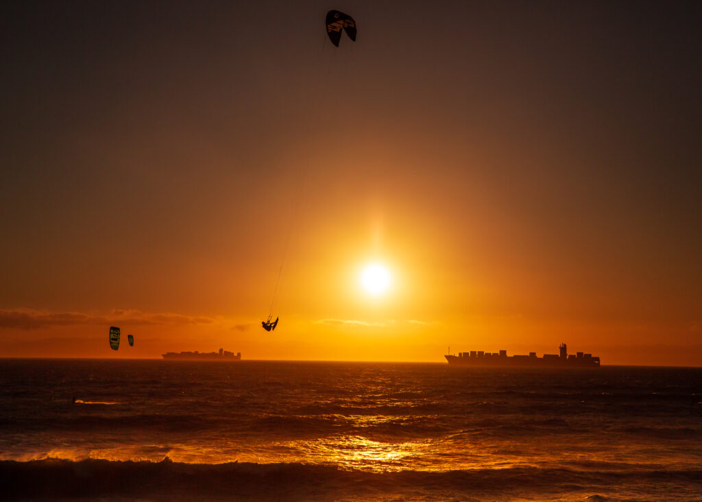 Kitesurfer silhouetted against a fiery sunset at Bloubergstrand, Cape Town, with cargo ships on the horizon.